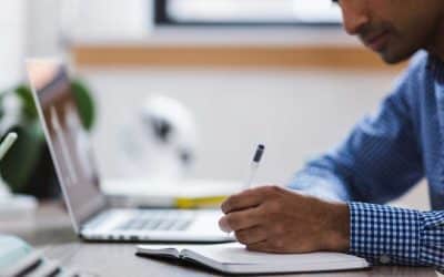 man writing in notebook with computer beside him.