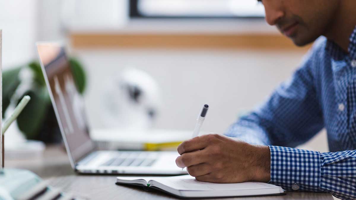 man writing in notebook with computer beside him.